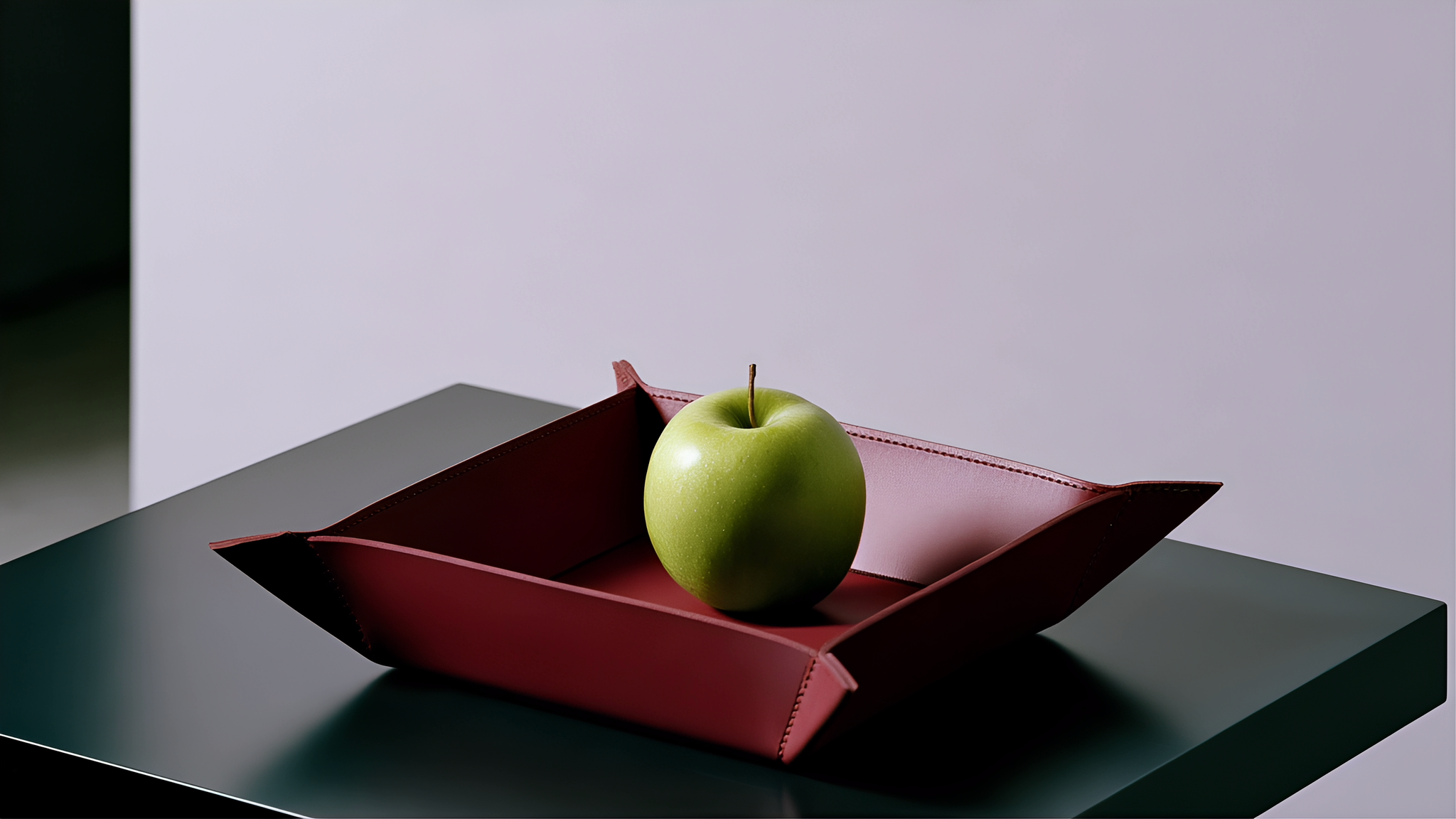 A green apple in an oxblood stitched leather tray against a pale lavender wall