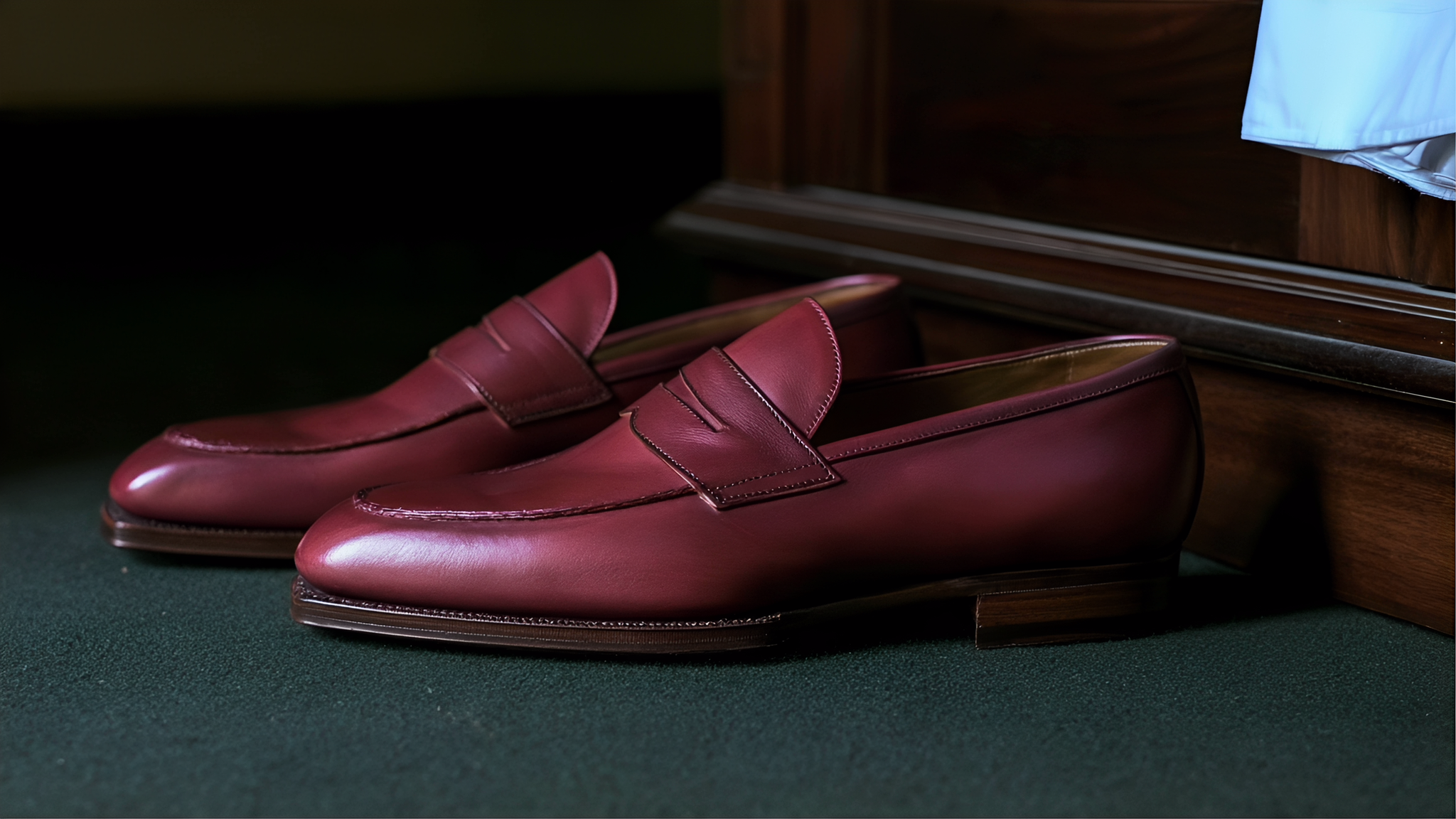 A footwear application concept: wine-red leather loafers on a dark green carpet beside a walnut wardrobe